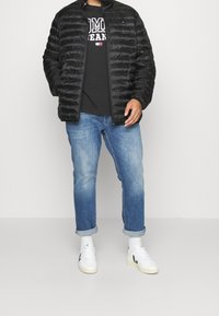 Schwarze Steppjacke mit einem gesteppten Muster, getragen über einem schwarzen Grafik-T-Shirt. Hellblaue Jeans mit umgekrempelten Manschetten und weiße Sneakers runden den Look ab.