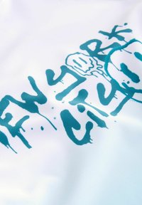 Vitt tyg med ett teal-färgat grafiskt tryck som inkluderar texten "NEW YORK CITY" i en droppande, stiliserad design.