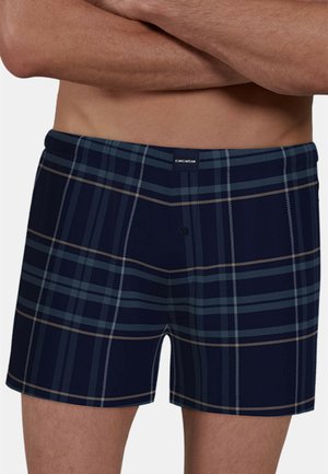 Male torso wearing navy blue and gray plaid boxer shorts with a button fly, arms crossed above the waist.