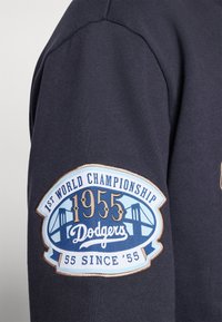 Molleton marine avec une manche présentant un patch brodé bleu et beige : "1er Championnat du Monde 1955 Dodgers 55 Depuis '55" avec un graphique de pont.