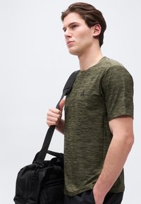 Νεαρός άνδρας με πράσινο ανάγλυφο μπλουζάκι που κουβαλάει μια μαύρη τσάντα duffel στον ώμο του, κοιτάζοντας μπροστά με φόντο απλό φόντο.