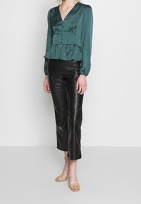 Blouse en satin teal avec un décolleté en V, un système de boutons et un détail peplum, associée à un pantalon droit en simili cuir noir et des sandales à brides nude.