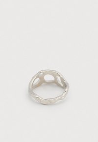 Ezüstszínű fém gyűrű szabálytalan áttört mintával és hullámos textúrájú felülettel, egyszínű világos háttér előtt bemutatva.