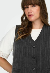 Grå rutvävd väst i textilmaterial med V-ringning, fyra svarta knappar och två sidofickor, som bärs över en vit skjorta.