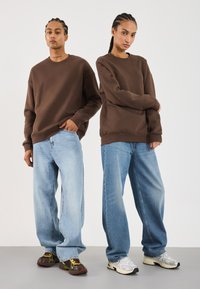 To modeller med flettehår har på seg brune hettegensere, løse lyseblå jeans og joggesko, og står foran en enkelt hvit bakgrunn.