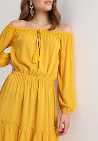 Gelbes schulterfreies Kleid mit elastischem Bund, langen Ärmeln und einem Frontverschluss mit einer Quaste. Strukturierter, leichter Stoff.