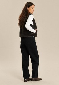 Vrouw met lang, krullend haar, die een zwart nepbont vest draagt over een witte blouse met lange mouwen, zwarte broek en bruine loafers, staand met handen in de zakken.