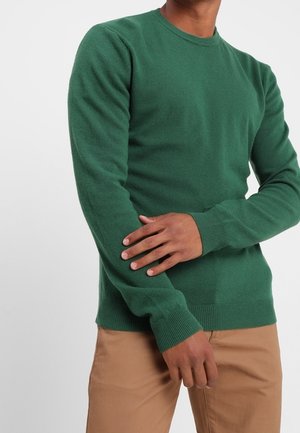 Mann trägt einen grünen Pullover mit Rundhalsausschnitt und langen Ärmeln sowie hellbraune Hose, eine Hand berührt den gegenüberliegenden Arm.