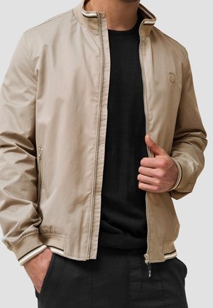 Mann trägt eine beige Bomberjacke mit Reißverschluss über einem schwarzen Hemd, eine Hand in der Tasche, die andere hält den Reißverschluss der Jacke.