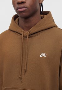 Κοντινό πλάνο ατόμου που φοράει καφέ φούτερ Nike SB με κορδόνια και μικρό λευκό λογότυπο στο στήθος, μονόχρωμο ανοιχτό γκρι φόντο.