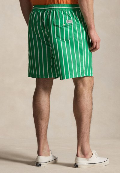 Vyras dėvi žalias maudymosi šortus su baltomis vertikaliomis juostelėmis ir mygtukais uždarytą užpakalinę kišenę, derina su baltais ilgais batais be raištelių.