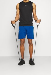 Musta ilmanhiha urheilupaita, jossa on valkoiset sivuraidat, siniset shortsit ja mustat lenkkarit. Malli pitää kädessään harjoitusvastusnauhoja.