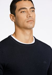 Granatowy sweter z okrągłym dekoltem, prążkowanymi mankietami i subtelnym logo. Gładka faktura, dopasowany krój, noszony na białej koszuli.