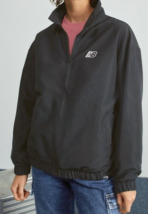 Personne portant une veste noire zippée avec un logo blanc sur la poitrine, un jean bleu et une chemise rose en dessous.