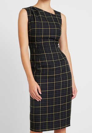 Robe noire ajustée sans manches avec un motif à carreaux blancs et jaunes, portée par une femme les bras détendus sur les côtés.