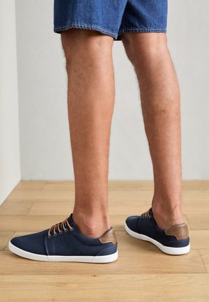 Marineblauwe canvas sneakers met bruine leren accenten, witte rubberen zolen en bruin veters, vanuit de zijkant weergegeven, gecombineerd met denim shorts.