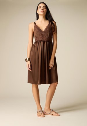 Młoda kobieta stojąca, z długimi ciemnymi włosami, ubrana w brązową koronkową sukienkę bez rękawów, złotą bransoletkę i zdobione sandały.