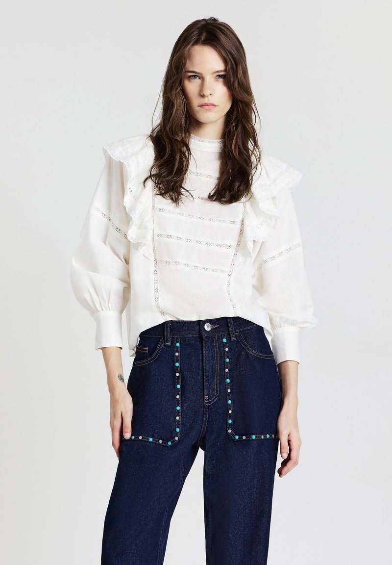 Blusa bianca con colletto arricciato e dettagli plissettati, abbinata a jeans in denim blu scuro con decorazioni colorate sulle tasche.