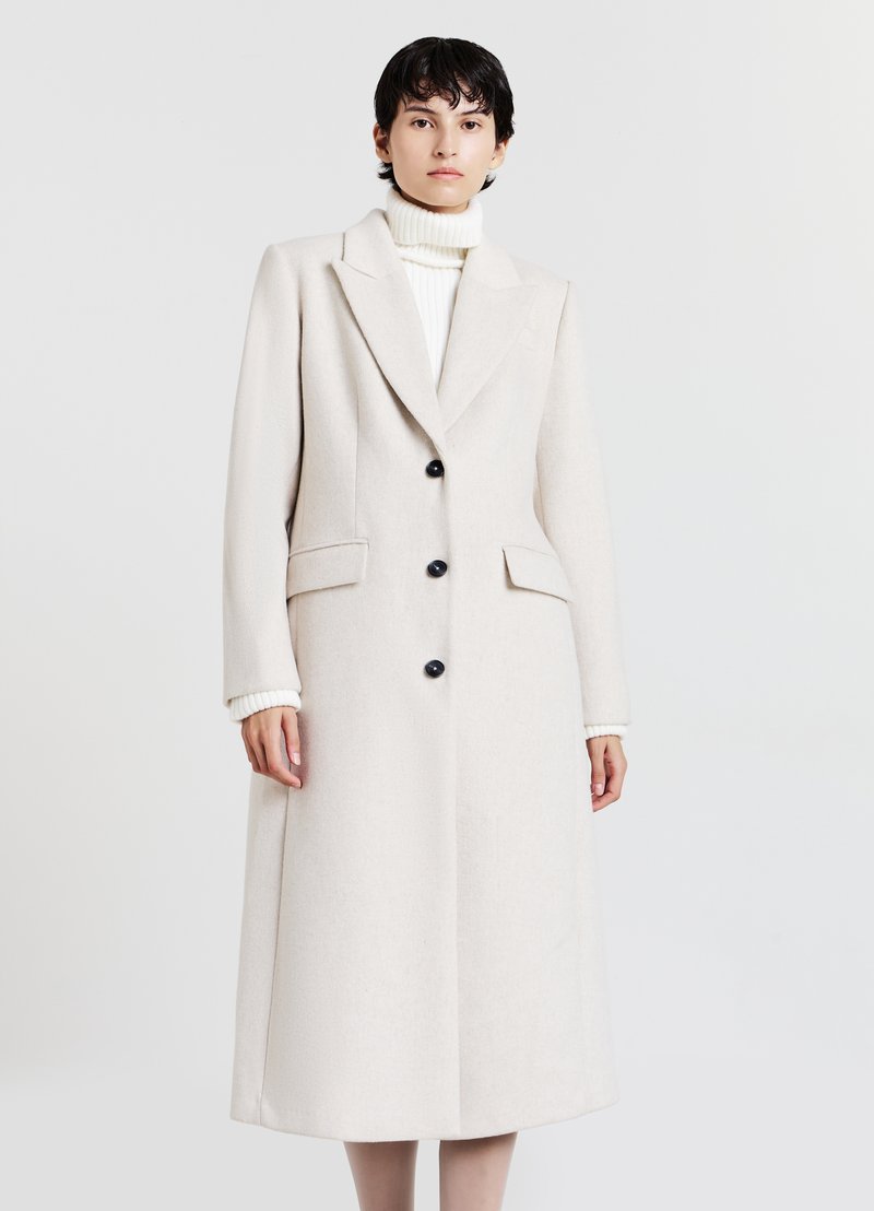 Cappotto lungo in lana beige chiaro con risvolti a punta, bottoni neri e tasche laterali. Indossato sopra un maglione a collo alto bianco a costine.