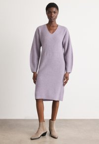 Lavandas krāsas ribota adījuma kleita ar V-veida kaklu, garām bumbveida piedurknēm un garumu līdz viduvēm. Valkāta kopā ar bēšiem potīšu zābakiem ar saliktu papēdi.