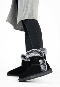 Las botas de tobillo de ante negro presentan un puño de piel sintética gris y un detalle de hebilla, con una suela de goma plana y una textura suave.