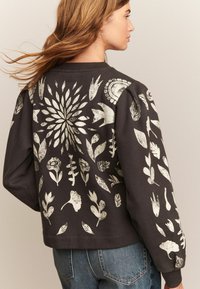 Kvinde set bagfra, iført en sort sweatshirt med hvide botaniske mønstre, parret med blå jeans, mod en neutral baggrund.