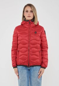 Donna con capelli biondi lisci, che indossa un giubbotto imbottito rosso con cappuccio e jeans azzurri chiari, in piedi davanti a uno sfondo bianco.