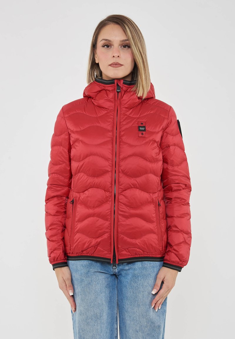 Donna con capelli biondi lisci, che indossa un giubbotto imbottito rosso con cappuccio e jeans azzurri chiari, in piedi davanti a uno sfondo bianco.
