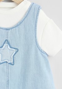 Farmernadrág ruha kék csillag hímzéssel, fehér bordázott pólóval párosítva. A ruha ujjatlan kialakítású, világoskék színű.