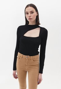 Sort langærmet top med et sideudskæring og ribbet tekstur, parret med beige tætsiddende bukser med forlommer og knaplukning.