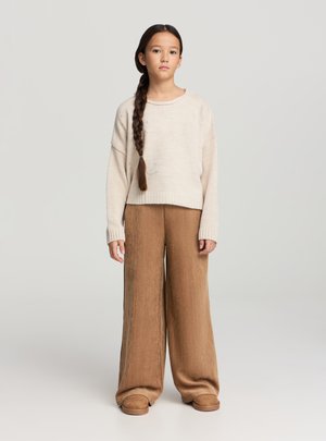 Ragazza con lunga treccia che indossa un maglione beige lavorato a maglia, pantaloni larghi marroni e pantofole marroni, in piedi contro uno sfondo chiaro e semplice.