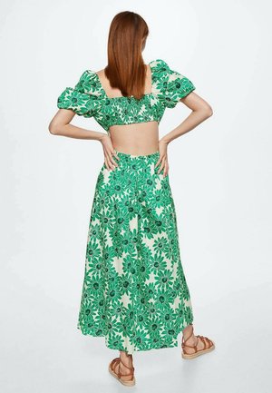 Kobieta o prostych brązowych włosach, ubrana w zielony kwiatowy krótki top z bufkami na rękawach oraz pasujące, szerokie spodnie z wysokim stanem, stojąca z rękami na biodrach.