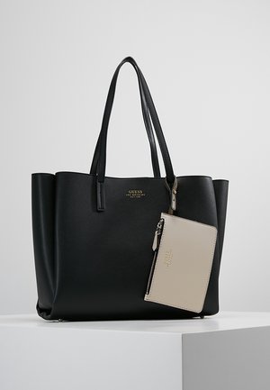 Zwarte Guess tote bag met twee handvatten en een beige zip-pouch eraan bevestigd, getoond op een wit voetstuk tegen een lichtgrijze achtergrond.