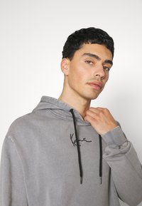 Graue Kapuzenpullover mit schwarzen Kordeln, ausgestattet mit einem gestickten Logo auf der Brust. Glatte Stofftextur mit lockerer Passform.