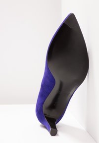 Chaussure à talon haut en daim violet unique avec un bout pointu incliné vers le haut, montrant une semelle noire lisse et un talon fin sur fond blanc.
