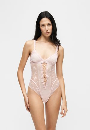 Woman with long dark hair wearing light pink lace bodysuit with crisscross detail on front, standing against plain light background.