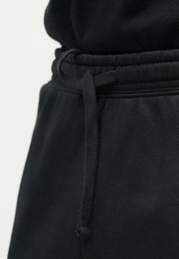 Gros plan sur la taille d'un pantalon de jogging noir avec une bande élastique et un détail de cordon noué, sur un fond clair.