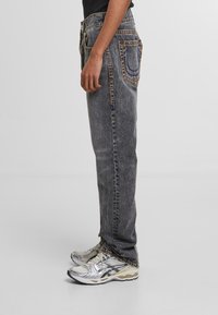 Grey denim jeans with a straight fit, featuring contrasting stitching, five pockets, and a mid-rise waist. Worn with silver athletic shoes.