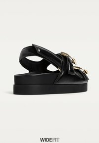 Sort lædersandal med en tyk sort platformssål, prydet med to guldspænder og et åbent bagdesign for nem påføring.