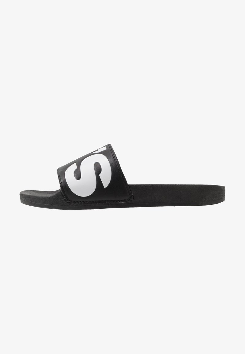 Tongs noires avec une large sangle présentant un logo blanc contrastant, semelle plate et semelle intérieure texturée pour une meilleure adhérence.