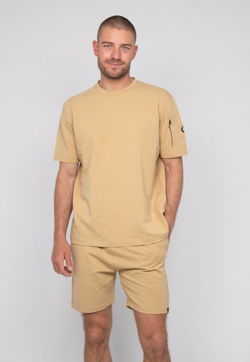 T-shirt en coton beige à manches courtes et coupe droite, associé à un short assorti. Le t-shirt présente une poche sur la manche.