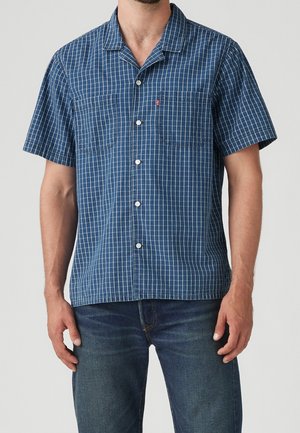 Man draagt een blauw, kort mouw knoophemd met witte verticale en horizontale strepen en blauwe jeans, staand tegen een effen achtergrond.