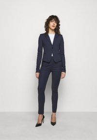 Tengerészkék, két részből álló öltöny, amely magában foglal egy egygombos, testre szabott zakót és slim-fit nadrágot, fekete magassarkú stiletto cipővel párosítva.