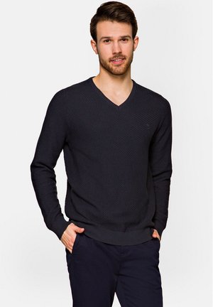 Ciemnoniebieski sweter w serek z teksturowanej tkaniny, z długimi rękawami i ściągaczami. Model pozuje z rękami w kieszeniach, nosząc ciemne spodnie.
