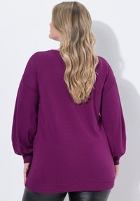 Donna con lunghi capelli biondi ondulati, che indossa una maglietta lunga viola a maniche lunghe e pantaloni neri, in piedi contro un muro bianco, di spalle rispetto all'osservatore.