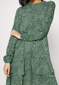 Vihreä kuvioitu mekko, joka on valmistettu kevyestä kankaasta. Mekossa on pitkät hihat, pyöreä pääntie ja kerroksellinen muotoilu, jossa on tekstuurisia yksityiskohtia.