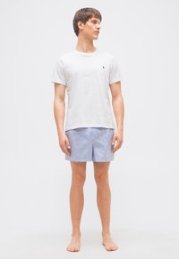 Νεαρός άνδρας στέκεται ξυπόλητος, φορώντας λευκό πουκάμισο κοντού μανικιού και μπλε-λευκή ριγέ βερμούδα, κοιτάζοντας προς τα αριστερά του.