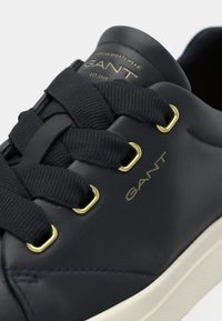 Σκούρο μπλε δερμάτινο αθλητικό παπούτσι GANT με φαρδιές μαύρες κορδόνια περασμένα από χρυσά οπτικά και λευκή λαστιχένια σόλα, με το λογότυπο ορατό στη γλώσσα και στο πλάι.