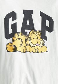 Bílý bavlněný tričko se designem kreslené kočky se šikmými oranžovými a žlutými pruhy, odpočívající pod tučným černým textem "GAP."