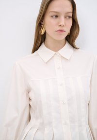 Lichtcrème knoopshirt van stof, met een gestructureerde kraag, verticale streepjes en geplooid detail aan de onderkant.